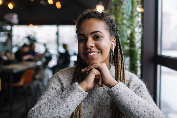 Портрет темнокожей женщины тысячелетия в наушниках Bluetooth прослушивания позитивной музыки подкаст во время отдыха в кафетерии, счастливое поколение Z в наушниках улыбаясь в камеру наслаждаясь аудио песни - Фото, изображение