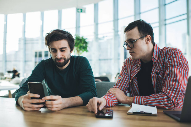 Content young male friends in casual clothes sitting at wooden table and browsing modern smartphone in creative workspace on daytime - Фото, зображення