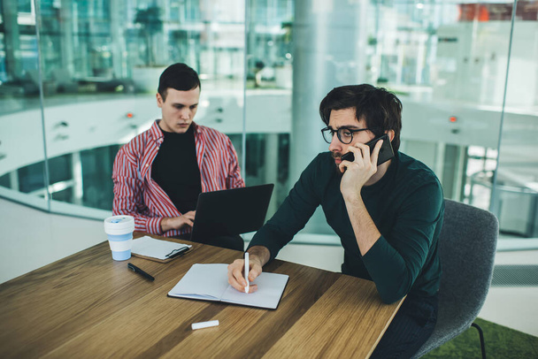 Busy male entrepreneur wearing casual outfit answering phone call and taking notes while sitting at table with concentrated coworker browsing netbook in spacious glass office - Photo, Image