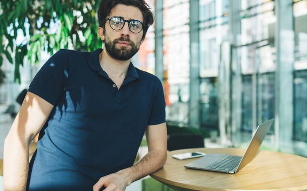 Focused bearded ethnic male freelancer in casual wear and glasses sitting at wooden table and looking at camera while having break - Fotoğraf, Görsel