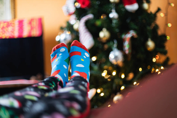 Nahaufnahme einer Person in lustigen Weihnachtssocken, die ihre Füße in der Nähe eines Weihnachtsbaums hochhält - Foto, Bild