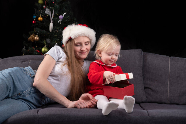 Η νεαρή μαμά με το καπέλο και το μωρό ανοίγει το χριστουγεννιάτικο δώρο σε μαύρο φόντο. Νέο έτος. - Φωτογραφία, εικόνα