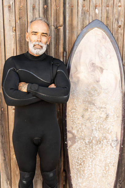A vertical shot of a happy mature male in a wetsuit with a surfing board - Foto, afbeelding