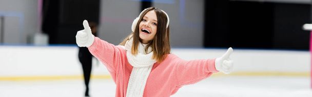 Ευτυχισμένη νεαρή γυναίκα με ωτοασπίδες και μαντήλι που δείχνει αντίχειρες στο παγοδρόμιο, πανό - Φωτογραφία, εικόνα