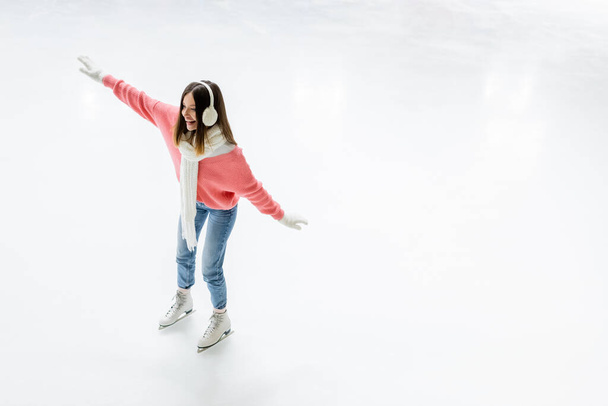 vista de ángulo alto de la joven feliz en orejeras y bufanda patinaje en pista de hielo - Foto, Imagen