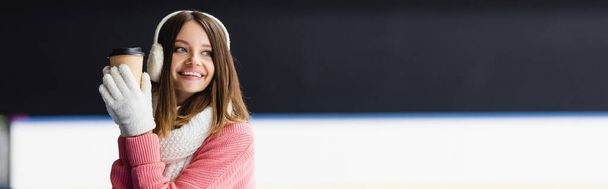 cheerful young woman in ear muffs and scarf holding paper cup on ice rink, banner - Photo, image