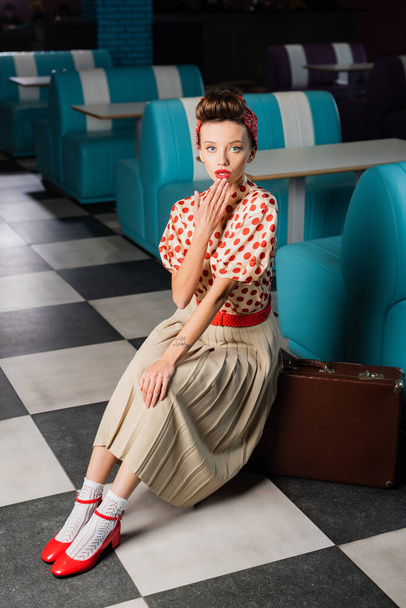 surprised pin up woman in dress sitting near retro suitcase in cafe - Fotoğraf, Görsel