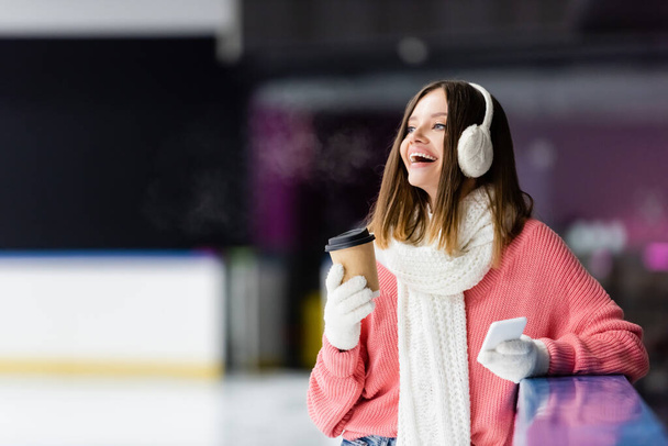 donna eccitata in paraorecchie tenendo tazza di carta e smartphone sulla pista di pattinaggio su ghiaccio  - Foto, immagini