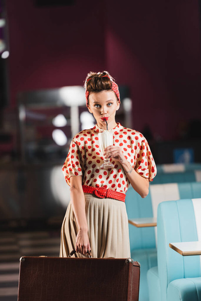 pin up woman holding suitcase and drinking tasty milkshake in cafe  - Photo, image