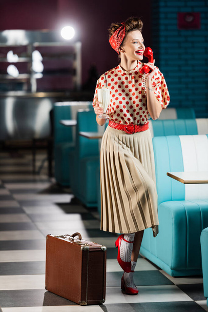 happy pin up woman talking on telephone and holding tasty milkshake near retro suitcase in cafe  - Фото, изображение
