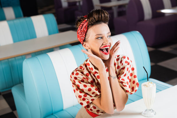 excited pin up woman sitting near milkshake on table  - Foto, Imagen