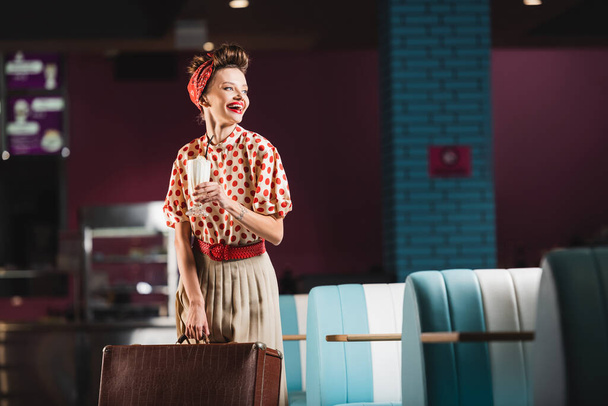 positive pin up woman holding suitcase and cocktail in cafe  - 写真・画像