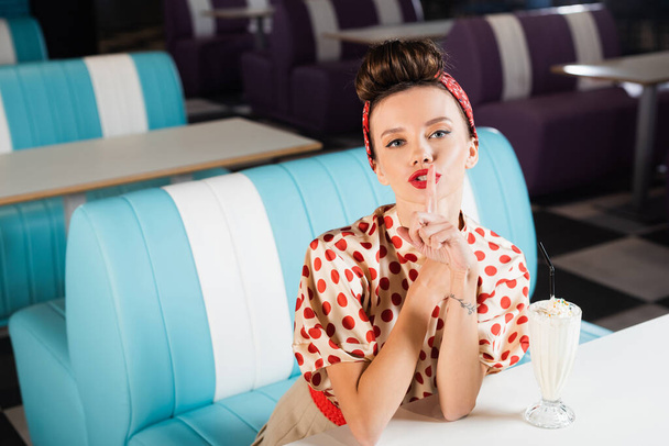 young pin up woman showing hush sign near milkshake on table  - Foto, Imagen