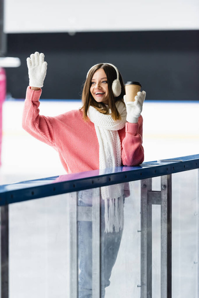 mujer excitada en orejeras y suéter rosa sosteniendo taza de papel y agitando la mano en la pista de hielo  - Foto, imagen