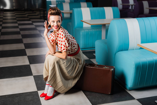 happy pin up woman sitting on vintage suitcase in cafe - Foto, afbeelding