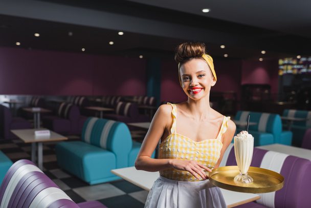 cheerful pin up waitress holding tray with milkshake in cafe  - Fotografie, Obrázek