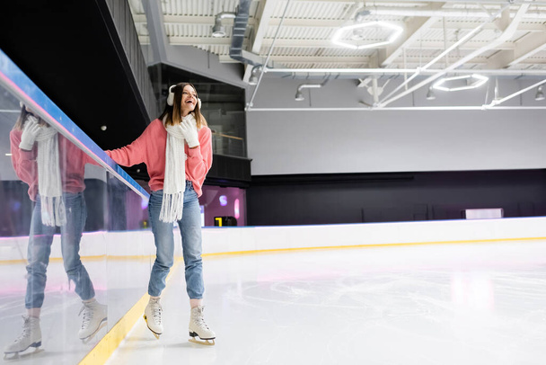 full length of positive young woman in white gloves and ear muffs and pink sweater skating on ice rink - Фото, изображение
