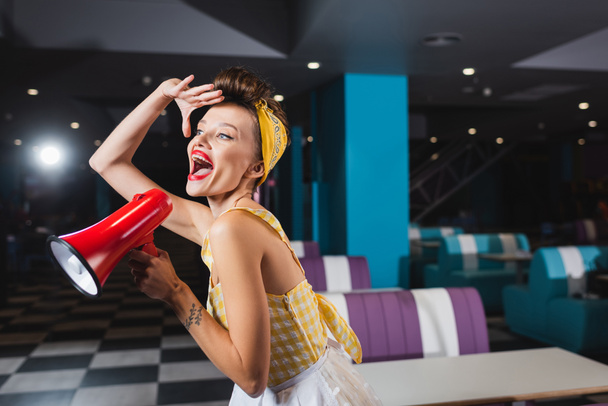 amazed pin up woman screaming in megaphone in retro cafe  - Fotografie, Obrázek