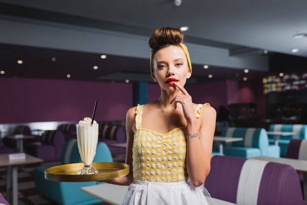 curious pin up waitress with tattoo holding tray and milkshake in cafe  - Foto, imagen