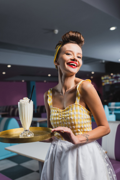 joyful pin up waitress with tattoo holding tray and tasty milkshake in cafe  - Foto, Imagem