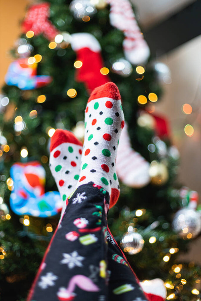 A vertical shot of a person in cozy pajamas and socks holding up their feet near a Christmas tree - Foto, afbeelding