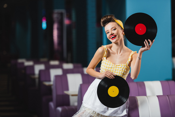 pin up woman with red lips holding retro vinyl records and smiling in cafe  - Fotó, kép