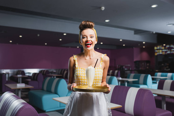 cheerful pin up waitress in dress holding tray with milkshake in cafe  - Foto, Bild