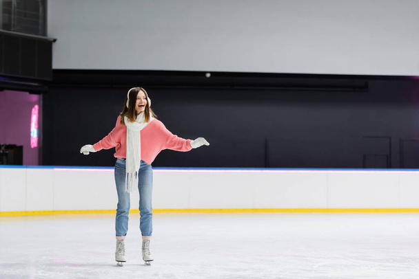full length of astonished woman in white scarf, sweater and ear muffs skating on ice rink - Photo, Image