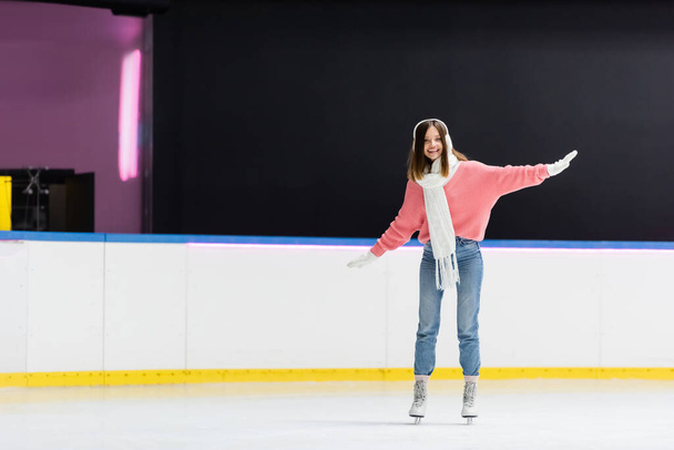 full length of smiling woman in ear muffs and winter outfit skating on ice rink - Photo, Image