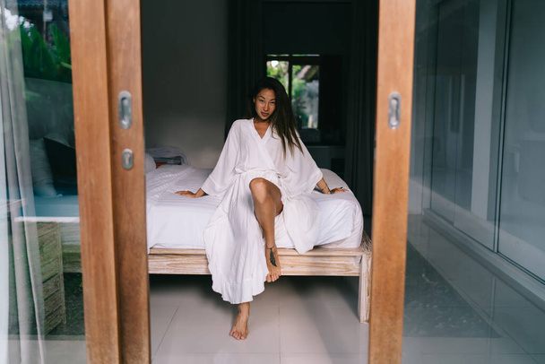 Full body barefoot sensual Asian woman in white dress sitting on comfortable bed in hotel room during vacation on Bali - Photo, Image