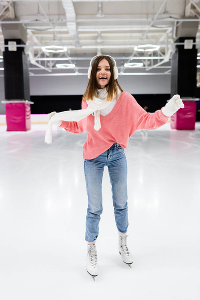 pleine longueur de femme heureuse dans des cache-oreilles et tenue d'hiver patinage et tenant écharpe sur la patinoire - Photo, image