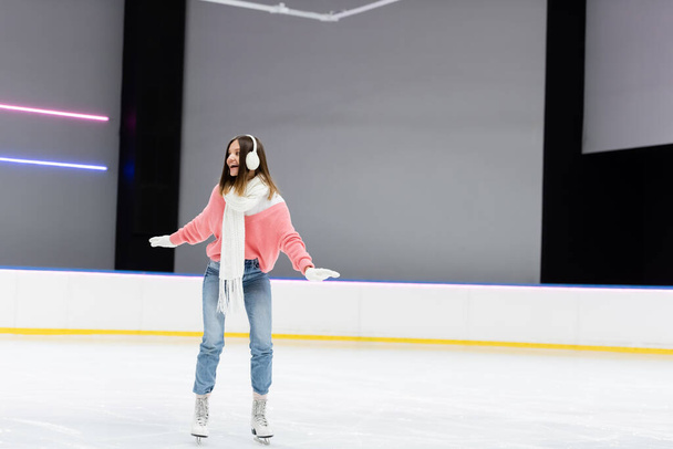 full length of happy woman in ear muffs and winter outfit skating on frozen ice rink  - Foto, Imagem