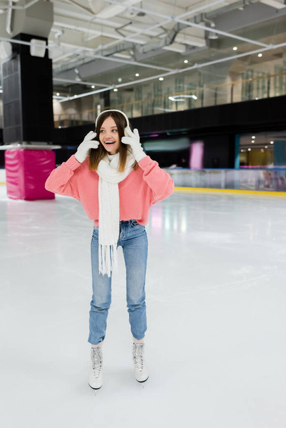 full length of cheerful young woman in white ear muffs and winter outfit skating on frozen ice rink  - Photo, image