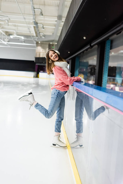 full length of happy woman in sweater, white scarf and ear muffs posing on ice rink - Foto, Bild