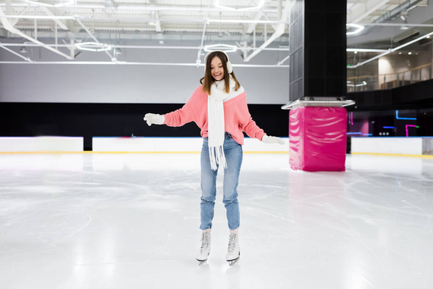 full length of happy woman in knitted sweater, ear muffs and winter outfit skating on ice rink  - Foto, Bild