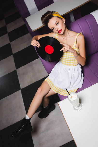 vista de ángulo alto de la mujer pin up con los labios rojos que sostienen el disco de vinilo en la cafetería  - Foto, Imagen