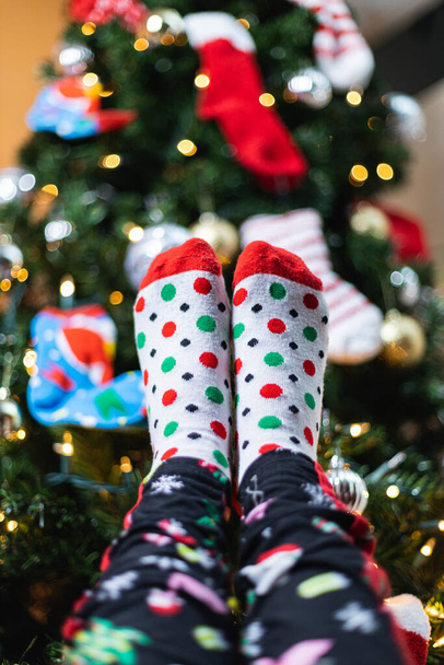 A vertical shot of a person holding up their feet in pajamas and socks near a Christmas tree - Fotó, kép