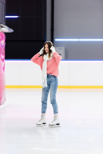 full length of cheerful woman in white scarf adjusting ear muffs and skating on ice rink - Photo, Image