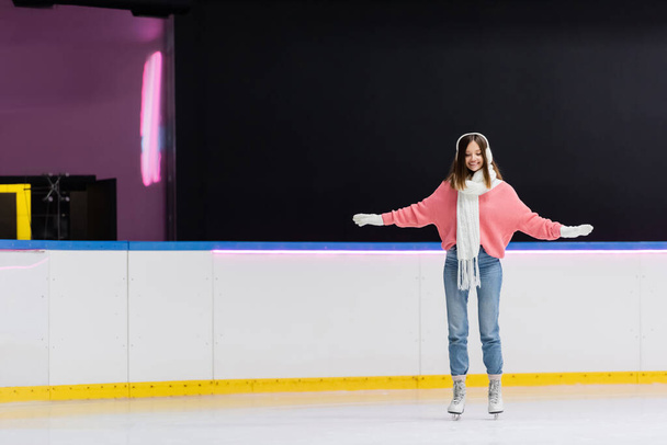 full length of cheerful woman in white ear muffs and knitted scarf skating with outstretched hands on ice rink - Фото, изображение