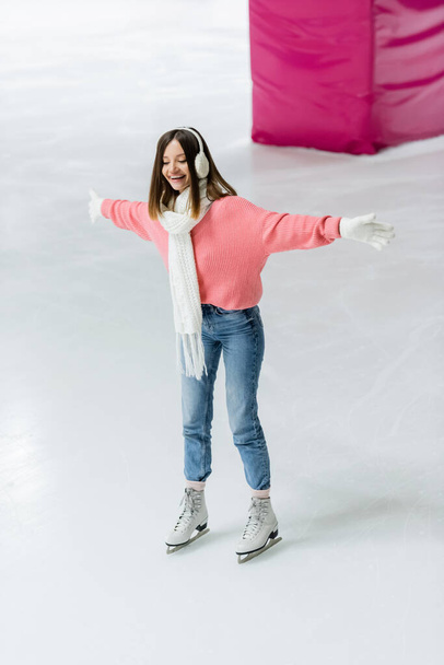 full length of joyful young woman in ear muffs and knitted scarf skating with outstretched hands on ice rink - Photo, image