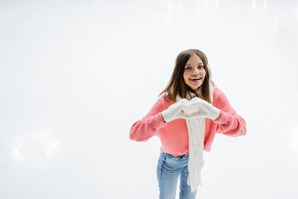 mujer joven feliz en orejeras blancas y bufanda de punto que muestra el signo del corazón en la pista de hielo - Foto, Imagen
