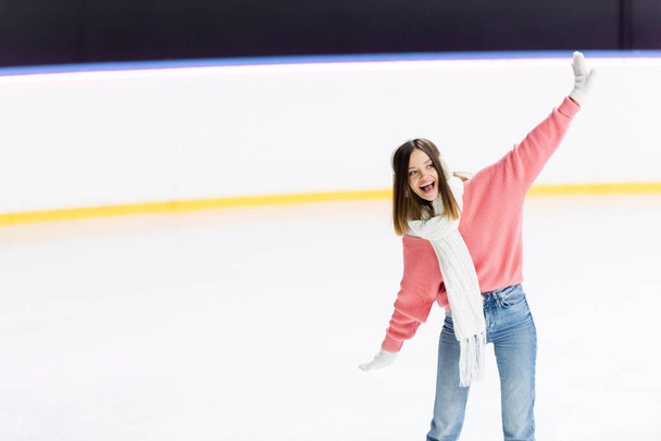 amazed young woman in white ear muffs and knitted scarf skating with outstretched hands on ice rink  - Φωτογραφία, εικόνα
