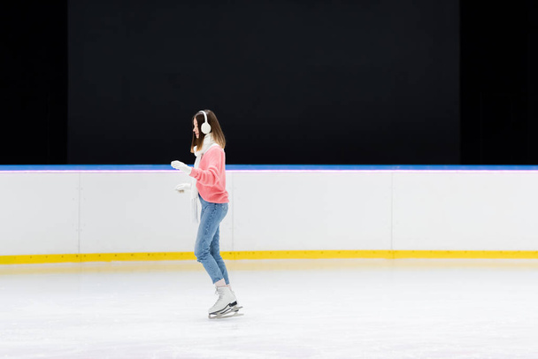 side view of young woman in white ear muffs skating on ice rink - Foto, afbeelding