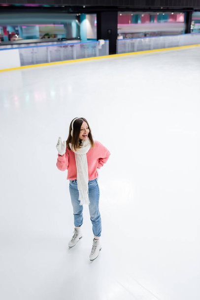 full length of happy woman posing with hand on hip and waving hand on ice rink  - Фото, изображение