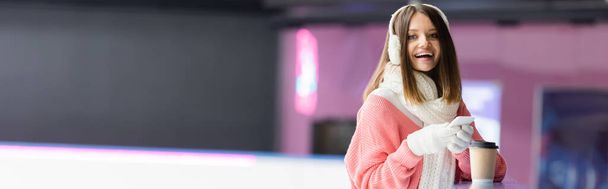 счастливая женщина в белых наушниках и розовый свитер держа сотовый телефон возле бумажной чашки, баннер  - Фото, изображение