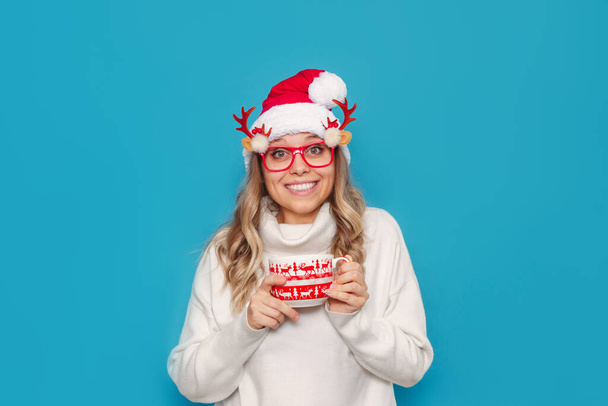 portrait of a happy girl in santa hat and glasses with mug on blue background - Foto, immagini