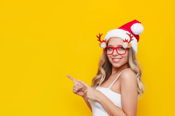 beautiful young woman in santa hat and glasses pointing up on yellow background - Фото, зображення