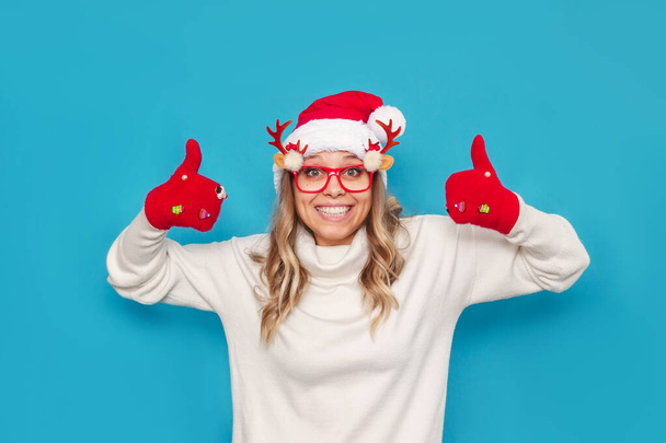 portrait of a young woman in santa claus hat and glasses showing thumbs up on blue background - 写真・画像