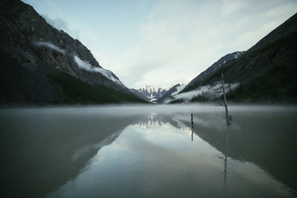 Scenic alpine landscape with snowy mountains in golden sunlight reflected on mirror mountain lake in fog among low clouds. Atmospheric highland scenery with low clouds on rocks and green mirror lake. - Photo, Image