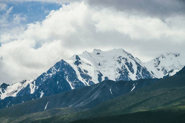 Φοβερό τοπίο βουνά με υψηλή χιονισμένη οροσειρά μεταξύ λευκά σύννεφα σε μπλε ουρανό. Ατμοσφαιρικό ορεινό τοπίο με χιόνι-λευκό μεγάλο βουνό κορυφογραμμή σε συννεφιασμένο καιρό. Υπέροχη χιονισμένη κορυφή - Φωτογραφία, εικόνα